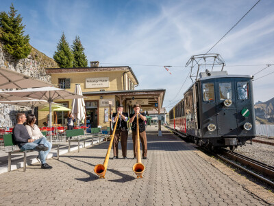 Schynige Platte Bahnhof Alphornblaeser zwei Personen auf Baenkli